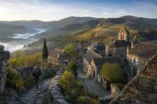 Quiet Cévennes village with schist walls, lauze roofs, morning light, vineyards, narrow cobbled streets, and a lone hiker.